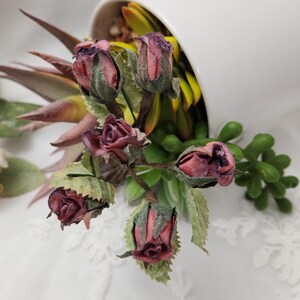 Pu&ograve; includere: Un contenitore di ceramica bianca rovesciato, che mostra un mix di piante grasse finte e boccioli di rosa secchi. I boccioli di rosa sono di un colore bordeaux intenso con foglie verdi. Le piante grasse sono di varie tonalit&agrave; di verde e giallo.
