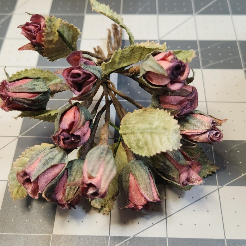 Pu&ograve; includere: Un mazzo di rose artificiali con petali rosa e bordeaux, foglie verdi e steli marroni. Le rose sono legate insieme con dello spago. I fiori sono disposti a grappolo, con alcuni boccioli ancora chiusi.