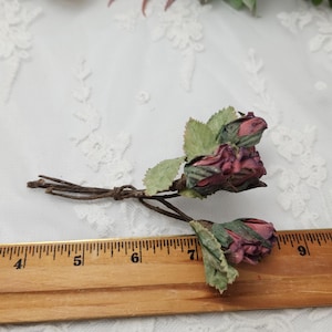 Pu&ograve; includere: Primo piano di due piccoli mazzi di rose artificiali. Le rose sono di un colore bordeaux intenso con foglie verdi. Gli steli sono marroni e legati insieme. Un righello di legno &egrave; in primo piano.