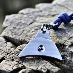May include: A silver-colored pendant with a unique, geometric design, featuring a central, circular accent. The pendant is suspended from a blue cord. The background is a blurred, natural setting with rocks.