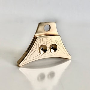 May include: A gold-colored metal whistle with a Celtic knot design and two holes. The whistle has a loop at the top for attaching a lanyard.