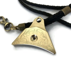 May include: A gold-colored whistle with an intricate floral design engraved on the surface. The whistle is attached to a black braided cord with a brass clasp.