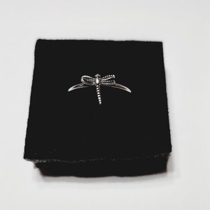 May include: A silver dragonfly ring. The ring showcases a detailed dragonfly design with intricate wings and a segmented body. The ring is presented on a black surface, set against a white background.