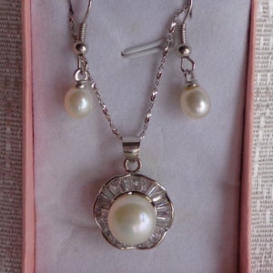 May include: A pearl necklace and earrings set in a pink box. The necklace has a silver pendant with a large pearl surrounded by small clear stones. The earrings are silver with small pearls.