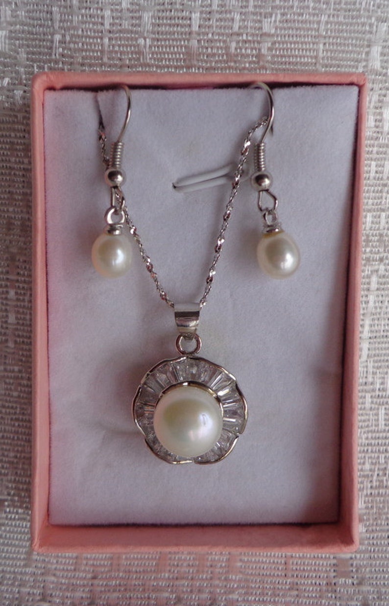 May include: A pearl necklace and earrings set in a pink box. The necklace has a silver pendant with a large pearl surrounded by small clear stones. The earrings are silver with small pearls.