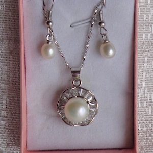 May include: A silver necklace and earrings set with a white pearl pendant surrounded by small clear stones. The pendant is in a flower shape.