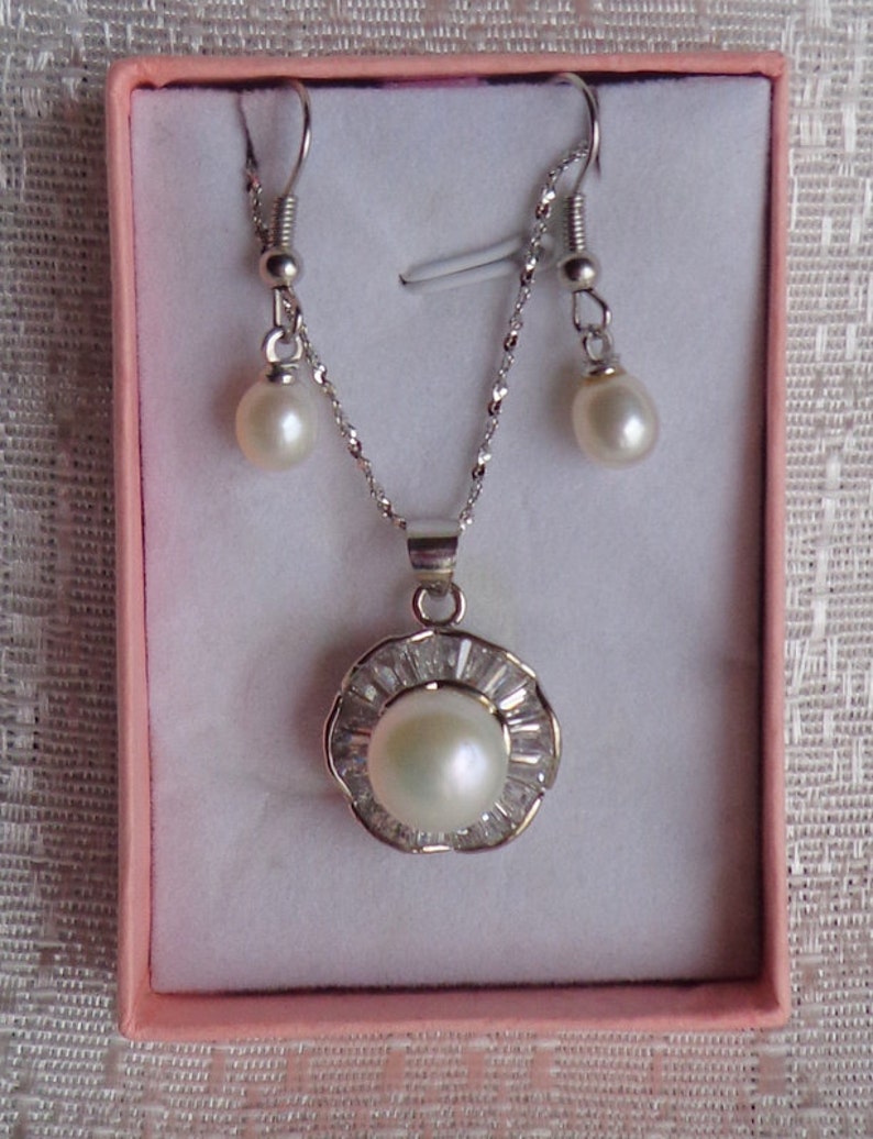 May include: A silver necklace and earrings set with a white pearl pendant surrounded by small clear stones. The pendant is in a flower shape.