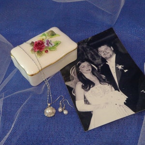May include: A white porcelain box with a floral design and a gold trim. The box is open and contains a silver necklace with a pearl pendant. There are also two silver earrings with pearl accents. The box is sitting on a blue background with a white veil.