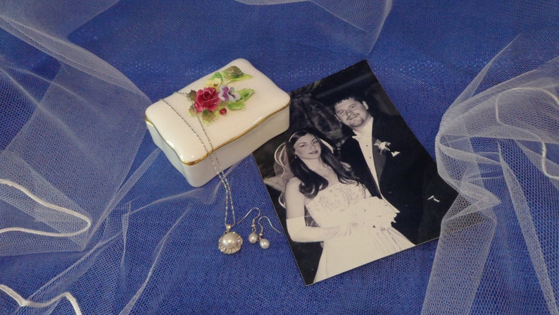 May include: A white porcelain box with a floral design and a gold trim. The box is open and contains a silver necklace with a pearl pendant. There are also two silver earrings with pearl accents. The box is sitting on a blue background with a white veil.