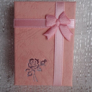 May include: A pink gift box with a satin ribbon and bow. The box has a silver embossed rose design.