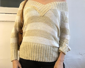 May include: Cream and beige striped off-the-shoulder sweater. The long-sleeved top features a V-neck and ribbed cuffs. The model is wearing black shorts and a brown shoulder bag.