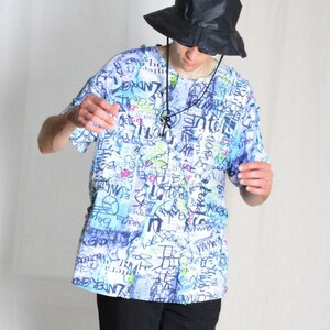 Può includere: Una persona indossa una camicia bianca e blu con stampa grafica, pantaloni neri e un cappello a secchiello nero. La camicia ha una varietà di testo colorato in stile graffiti stampato su di essa.