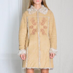May include: A long, tan suede coat with a faux fur collar and trim. The coat has a zipper closure and features embroidered paisley patterns in a reddish-brown color.
