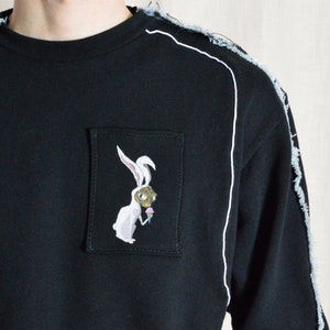 Puede incluir: Sudadera negra con un gr&aacute;fico de conejo blanco en un bolsillo de parche. La sudadera tiene costuras blancas a lo largo de las costuras y un aspecto desgastado.