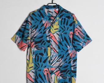 Op de afbeelding: Een overhemd met korte mouwen en knoopsluiting, voorzien van een levendig abstract patroon in tinten blauw, zwart, geel en koraalroze. Het kledingstuk heeft een klassieke kraag, een knoopsluiting aan de voorzijde en hangt aan een zwarte kledinghanger.