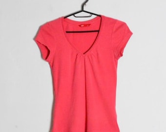 Op de afbeelding: Een fel koraalroze T-shirt met korte mouwen, een V-hals en een subtiel geplooid detail in het midden van de borst. Het shirt heeft een klein rood merklabel aan de linkeronderzoom en hangt aan een zwarte plastic kledinghanger.