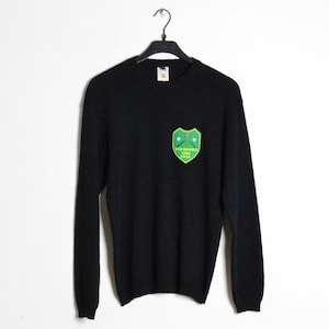 Puede incluir: Jersey negro de manga larga con un parche verde y amarillo en el pecho. El parche presenta un diseño de escudo con palos de golf cruzados y texto. El jersey está colgado de una percha negra sobre un fondo blanco.