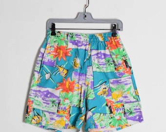 Op de afbeelding: Een korte broek met een levendige, meerkleurige tropische print op een heldere turquoise achtergrond. Het patroon bevat illustraties van oranje bloemen, groene palmbomen en gele vissen, tegen een achtergrond van paarse en witte accenten.