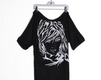 Op de afbeelding: Zwart shirt met korte mouwen en een witte afbeelding van een vrouwengezicht. Het ontwerp is een gestileerd portret met vloeiend haar en gezichtsdetails. Het shirt hangt aan een grijze hanger tegen een witte achtergrond.
