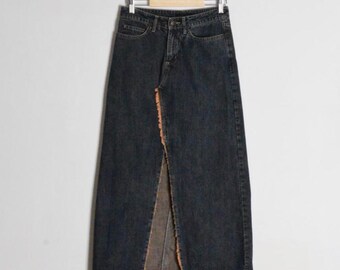 Op de afbeelding: Een lange rok van donker gewassen denim met een hoge taille, knoop- en ritssluiting, vijf zakken en een opvallende verticale split aan de voorzijde met rafelige randen die de lichtere binnenstof van het denim materiaal blootleggen.