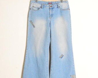 Op de afbeelding: Lichtblauwe denim jeans met een wijde pijp, hangend aan een houten hanger. De jeans hebben een vervaagde wassing en patches op de voorkant. Een klassieke, casual stijl.