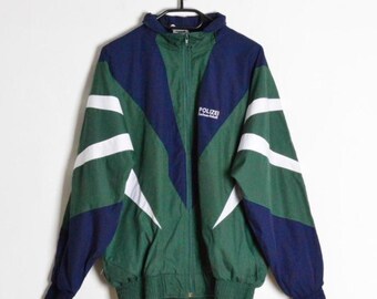 Op de afbeelding: Een groene, marineblauwe en witte vintage jas met het woord "POLIZEI" op de borst. De jas heeft een volledige ritssluiting, een hoge kraag en witte schuine strepen op de mouwen. De jas hangt aan een zwarte hanger.