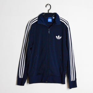 Può includere: Giacca da tuta blu navy con strisce bianche sulle maniche e logo bianco sul petto. La giacca ha una zip intera e un colletto rialzato. Classico capo di abbigliamento sportivo.