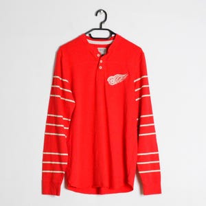 May include: A bright red long-sleeve henley shirt featuring a white winged wheel logo on the left chest. The sleeves have horizontal white stripes, and the neckline includes a two-button placket. The shirt is displayed on a black hanger.