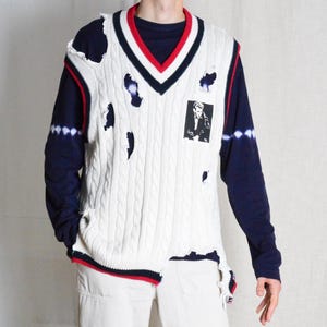 May include: A distressed white cable knit sweater vest with navy and red trim, worn over a navy long-sleeve shirt and paired with off-white cargo trousers. The vest features a black and white graphic and distressed holes.