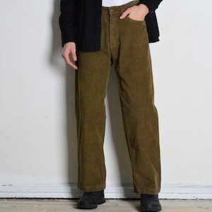 Pode incluir: Uma pessoa vestindo uma camisa preta sobre uma camiseta branca e calças de veludo cotelê verde oliva. A roupa é casual e as calças têm um corte reto.