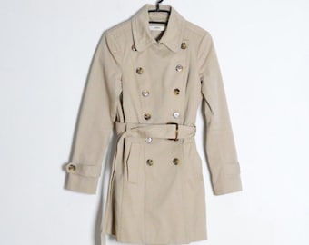 Op de afbeelding: Een beige dubbelrijige trenchcoat met een brede kraag, lange mouwen met knoopmanchetten en een bijpassende ceintuur met gesp. De jas is voorzien van twee verticale rijen bruine knopen en heeft twee voorzakken.