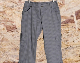 Op de afbeelding: Grijze capribroek met verticale krijtstrepen, met cargozakken en knoopdetails. De broek hangt aan een houten kleerhanger tegen een lichtbruine houten achtergrond. De capribroek is een casual stijl.