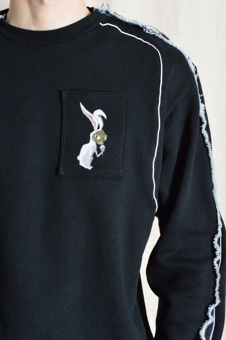 Puede incluir: Sudadera negra con un gr&aacute;fico de conejo blanco en un bolsillo de parche. La sudadera tiene costuras blancas a lo largo de las costuras y un aspecto desgastado.