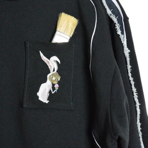 Puede incluir: Sudadera negra con ribete blanco y un detalle de denim desgastado en la manga. La sudadera tiene un bolsillo con un conejo blanco que lleva una m&aacute;scara de gato y sostiene un cono de helado bordado en &eacute;l. Un pincel sobresale del bolsillo.