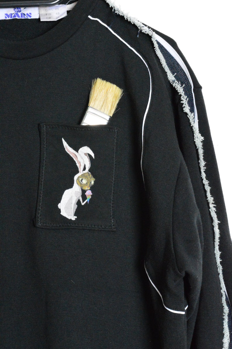 Puede incluir: Sudadera negra con ribete blanco y un detalle de denim desgastado en la manga. La sudadera tiene un bolsillo con un conejo blanco que lleva una m&aacute;scara de gato y sostiene un cono de helado bordado en &eacute;l. Un pincel sobresale del bolsillo.