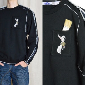 Puede incluir: Sudadera negra con ribetes blancos y un gr&aacute;fico de un conejo blanco en la parte delantera. El conejo sostiene un pincel y un cono de helado. La sudadera tiene un bolsillo en la parte delantera con un pincel que sobresale.