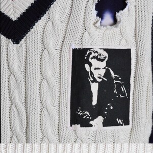 May include: Cream cable knit sweater vest with a V-neck featuring red, white, and navy blue stripes. A black and white graphic of a man smoking is on the front. The sweater has a distressed hole and a "MEDIUM" tag.