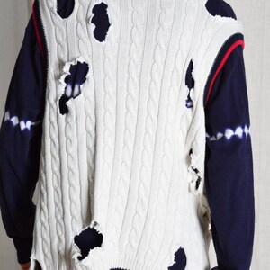 May include: White cable knit sweater vest with distressed holes, red and navy trim, and a navy blue long-sleeve shirt underneath. The vest has a classic cable knit pattern and a V-neck design.
