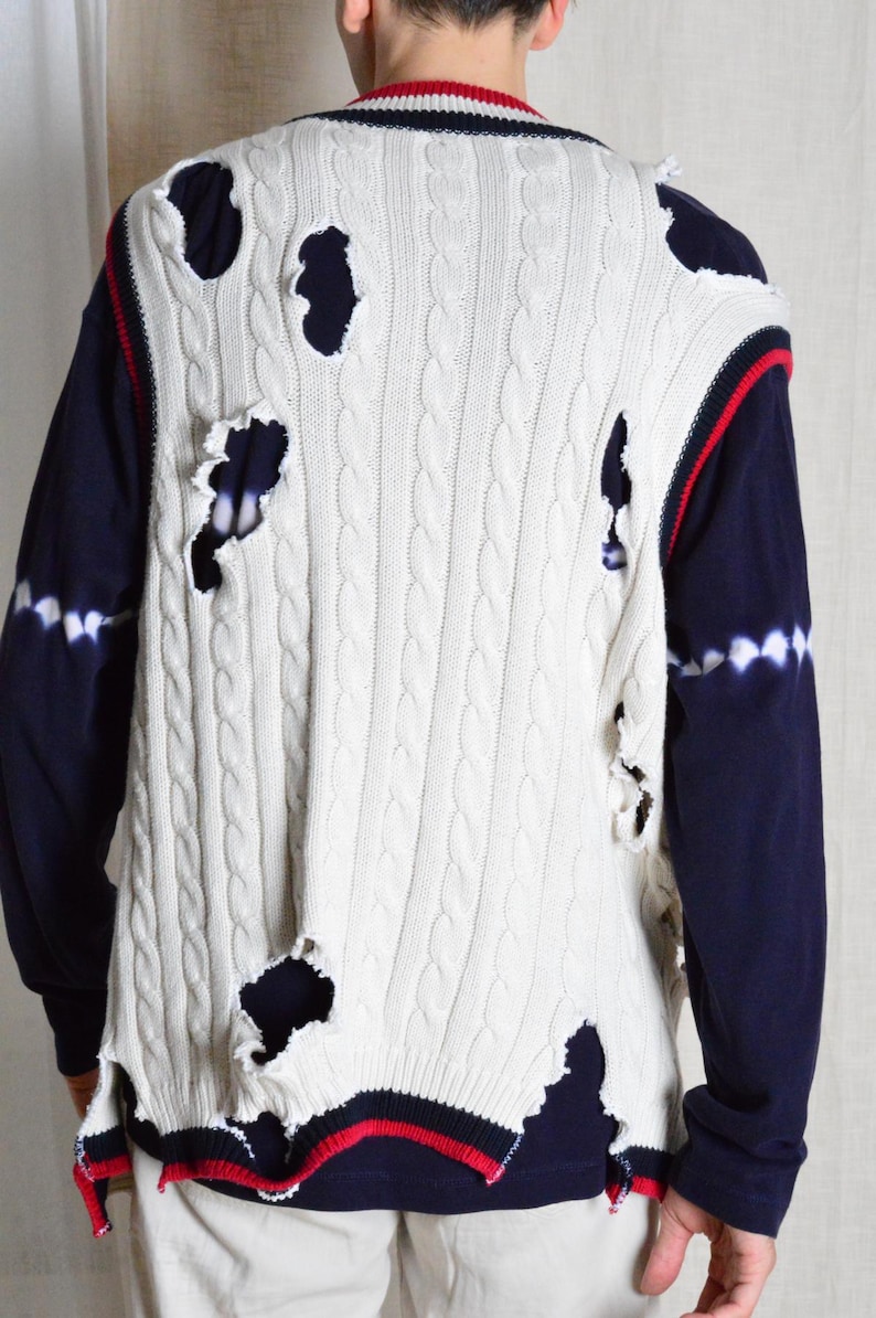 May include: White cable knit sweater vest with distressed holes, red and navy trim, and a navy blue long-sleeve shirt underneath. The vest has a classic cable knit pattern and a V-neck design.
