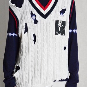 May include: White cable knit sweater vest with distressed holes, a V-neck, and red and navy stripes. A black and white image is on the front. Worn over a navy long-sleeve shirt.