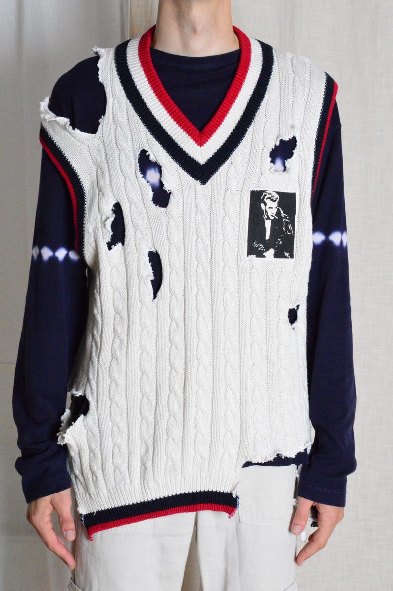 May include: White cable knit sweater vest with distressed holes, a V-neck, and red and navy stripes. A black and white image is on the front. Worn over a navy long-sleeve shirt.
