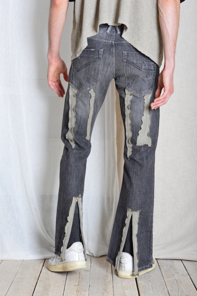 K&ouml;nnte beinhalten: Dunkelgraue Jeans mit Used-Details. Die Jeans hat vertikale, strukturierte Stoffakzente und Schlitze an den Kn&ouml;cheln. Wei&szlig;e Turnschuhe sind am unteren Ende der Jeans zu sehen.