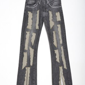 K&ouml;nnte beinhalten: Dunkelgraue Jeans mit zerfetzten, vertikalen, beigefarbenen Stoffakzenten. Die Jeans hat ein klassisches F&uuml;nf-Taschen-Design und ein gerades Bein mit einem Schlitz am Saum. Die Jeans h&auml;ngt an einem Holzb&uuml;gel vor wei&szlig;em Hintergrund.
