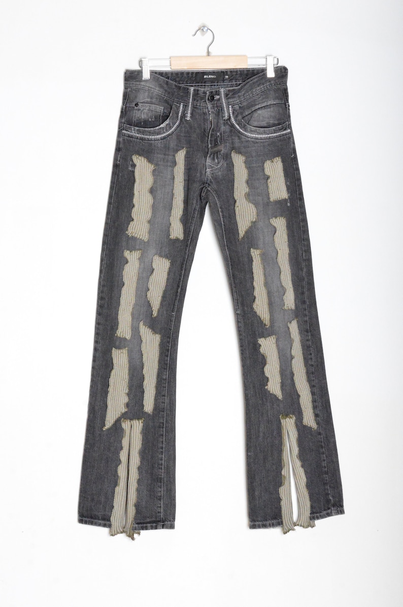 K&ouml;nnte beinhalten: Dunkelgraue Jeans mit zerfetzten, vertikalen, beigefarbenen Stoffakzenten. Die Jeans hat ein klassisches F&uuml;nf-Taschen-Design und ein gerades Bein mit einem Schlitz am Saum. Die Jeans h&auml;ngt an einem Holzb&uuml;gel vor wei&szlig;em Hintergrund.