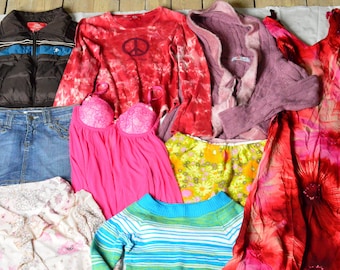 Op de afbeelding: Een verzameling kledingstukken, waaronder een spijkerrok, een roze en wit bloemetjesprint top, een blauw en wit gestreepte trui, een roze en wit bloemetjesprint jurk, een rood en wit tie-dye longsleeve shirt, een bruin puffer vest, een roze cardigan, een geel en groene bloemetjesprint rok en een rood en roze bloemetjesprint jurk.