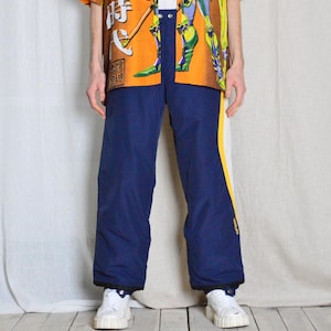 Może przedstawiać: Niebieskie spodnie nylonowe z żółtymi i białymi paskami po bokach. Spodnie mają sznurek w pasie i luźny krój.