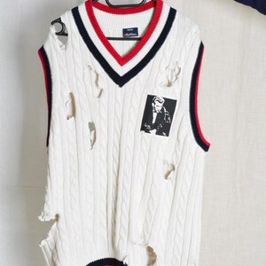 May include: Cream cable knit sweater vest with a V-neck, featuring red and navy blue stripes. The vest has distressed holes and a small black and white graphic patch. The vest is hanging on a black hanger.