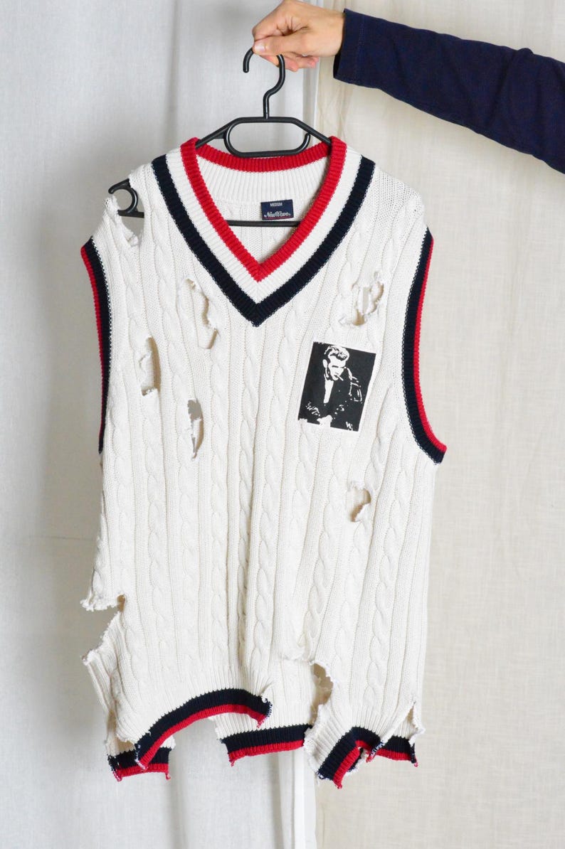 May include: Cream cable knit sweater vest with a V-neck, featuring red and navy blue stripes. The vest has distressed holes and a small black and white graphic patch. The vest is hanging on a black hanger.