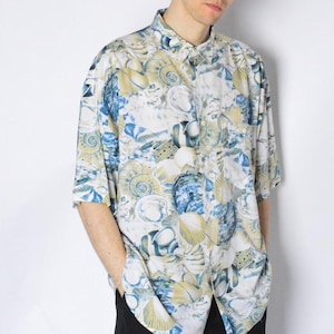 以下が含まれることがあります： カラフルな貝殻柄の白い半袖ボタンダウンシャツ。ゆったりとしたフィット感と襟があります。