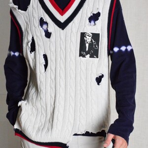 May include: Cream cable knit sweater vest with a V-neck, red and navy trim, and distressed holes. A black and white image is on the front. The model is wearing a navy blue long sleeve shirt. The vest is a unique fashion item.
